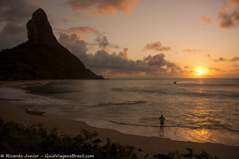 Imagem de uma pessoa nas areias com braços na cintura admirando o fim do por do sol.